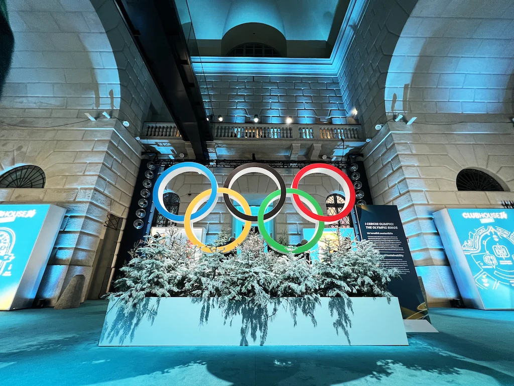 Olympic rings above a planter of snow-covered firs in a stone hall washed in icy blue light – Clubhouse 26 Milano, Italy, 2026 – hospitality design