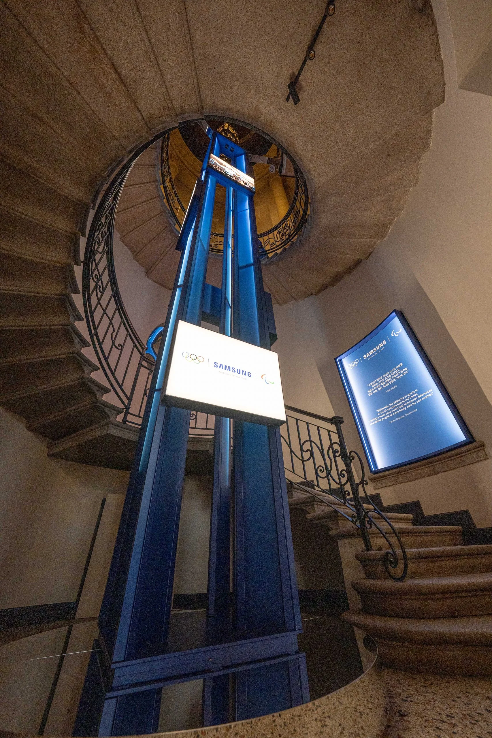 Blue steel lift shaft with Samsung and Olympic signage rising through a spiral stone staircase – Samsung House at Palazzo Serbelloni, Milan, Italy, 2026 – interior design