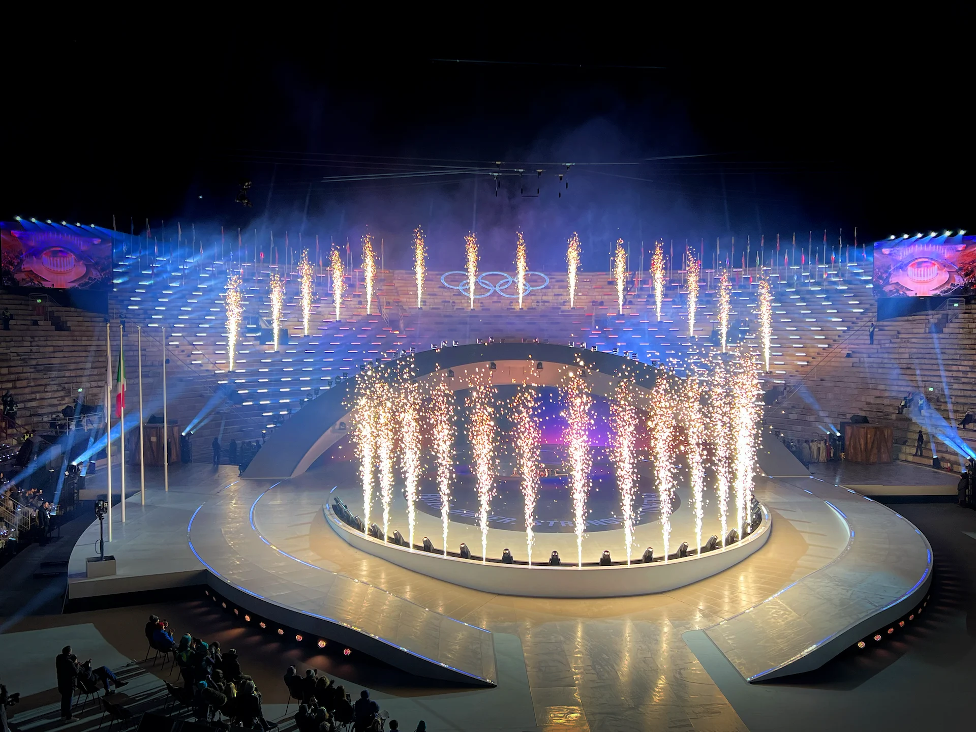 Fountains of sparks along the central stage and the arena steps beneath the Olympic rings – Milano Cortina 2026 Olympic Closing Ceremony, Verona, Italy – stage design