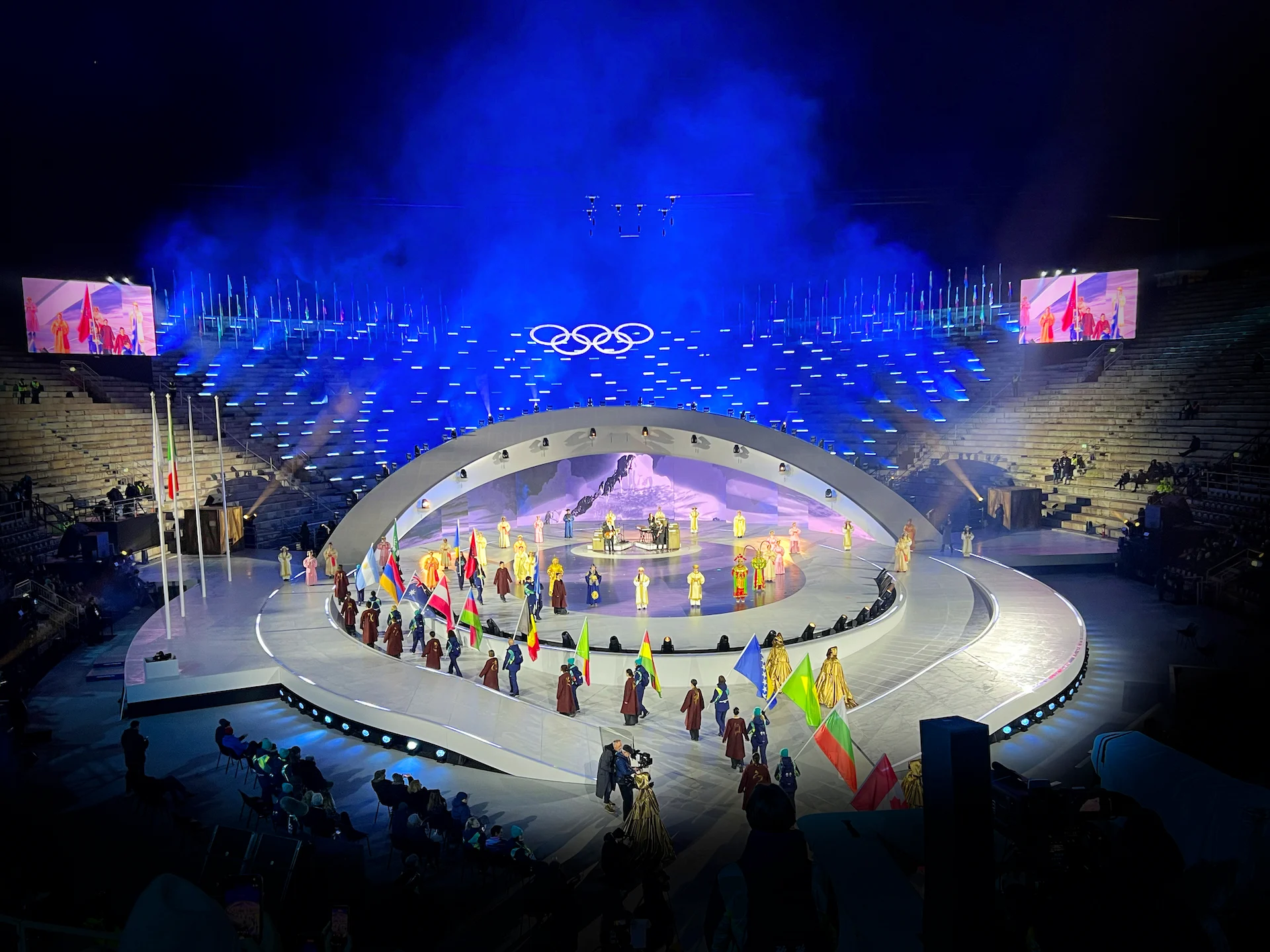 Flag bearers parading around the oval stage under the Olympic rings, blue haze over the arena steps – Milano Cortina 2026 Olympic Closing Ceremony, Verona, Italy – stage design