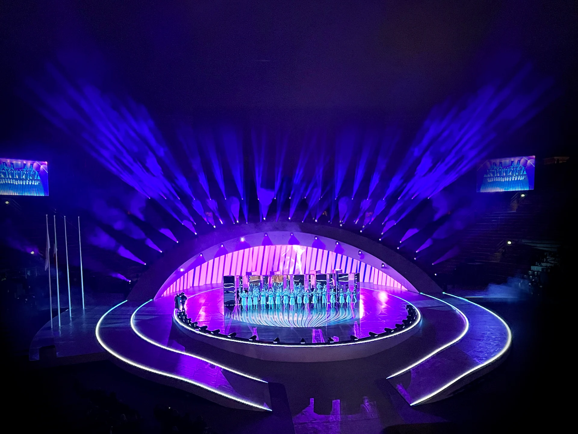 Choir on the central stage under the curved arch, framed by a fan of purple light beams – Milano Cortina 2026 Olympic Closing Ceremony, Verona, Italy – stage design