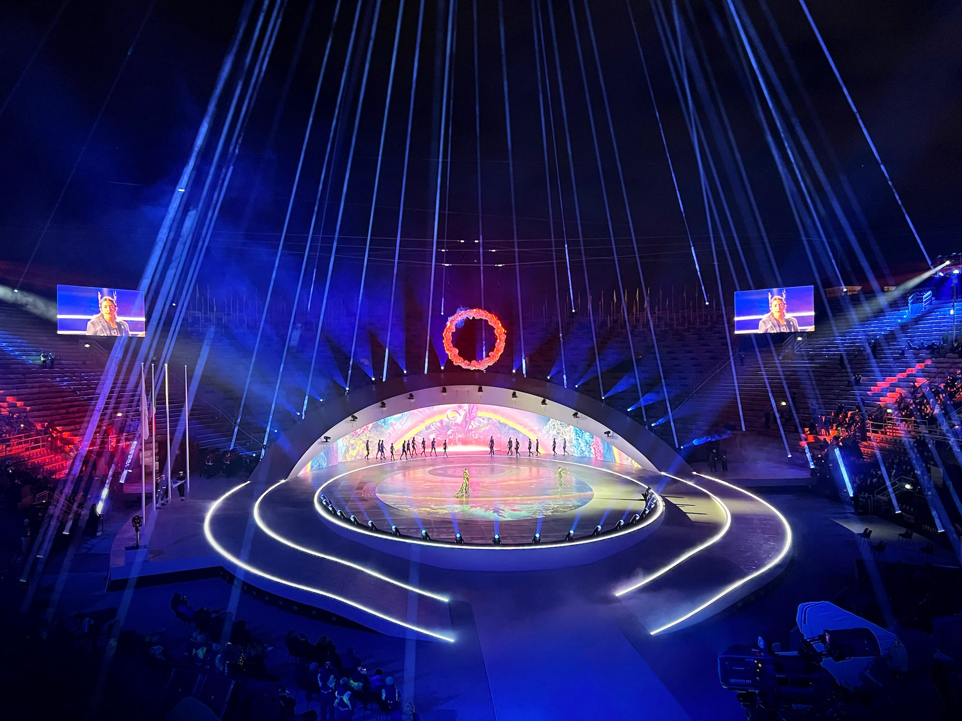 Performers on the illuminated stage under a ring of flames, with light beams falling from above – Milano Cortina 2026 Olympic Closing Ceremony, Verona, Italy – stage design