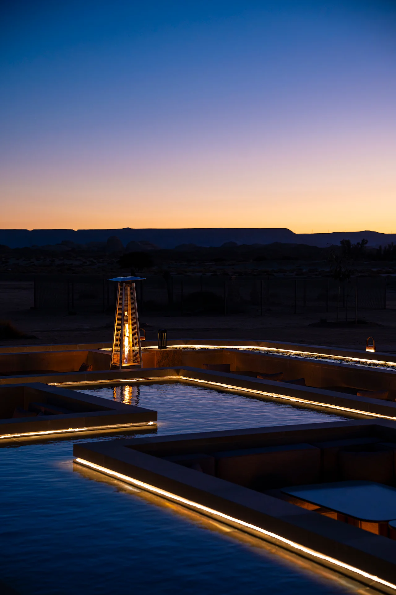 Glowing patio heater and lit basin edges at twilight, with desert mesas on the horizon – Sky View Restaurant at The Chedi Hegra, AlUla, Saudi Arabia, 2026 – architecture