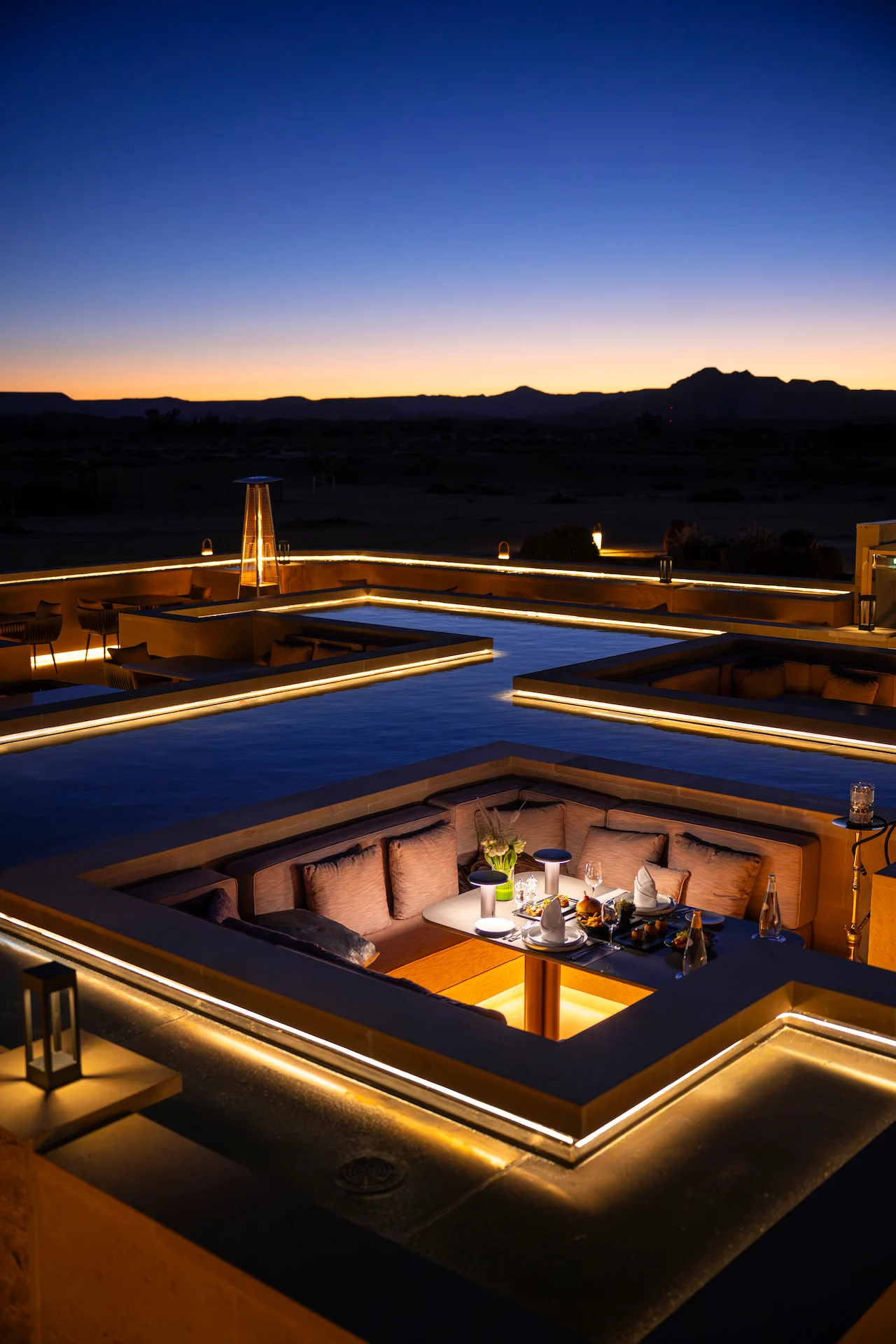 Sunken booth with a table set for dinner at blue hour, light lines tracing the basin edges – Sky View Restaurant at The Chedi Hegra, AlUla, Saudi Arabia, 2026 – architecture