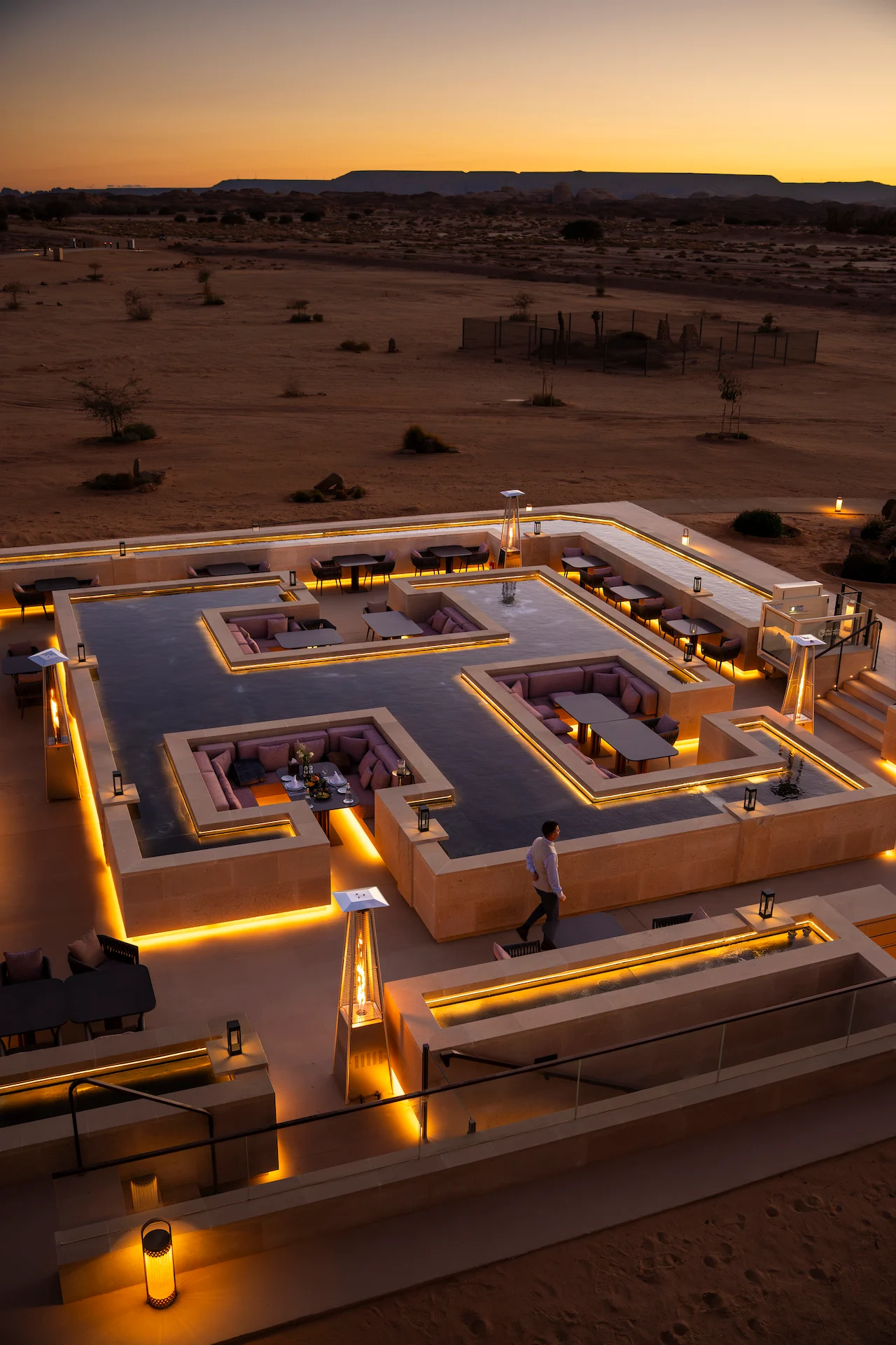 Elevated view of the restaurant terrace at sunset, a waiter walking past patio heaters, desert plain behind – Sky View Restaurant at The Chedi Hegra, AlUla, Saudi Arabia, 2026 – architecture