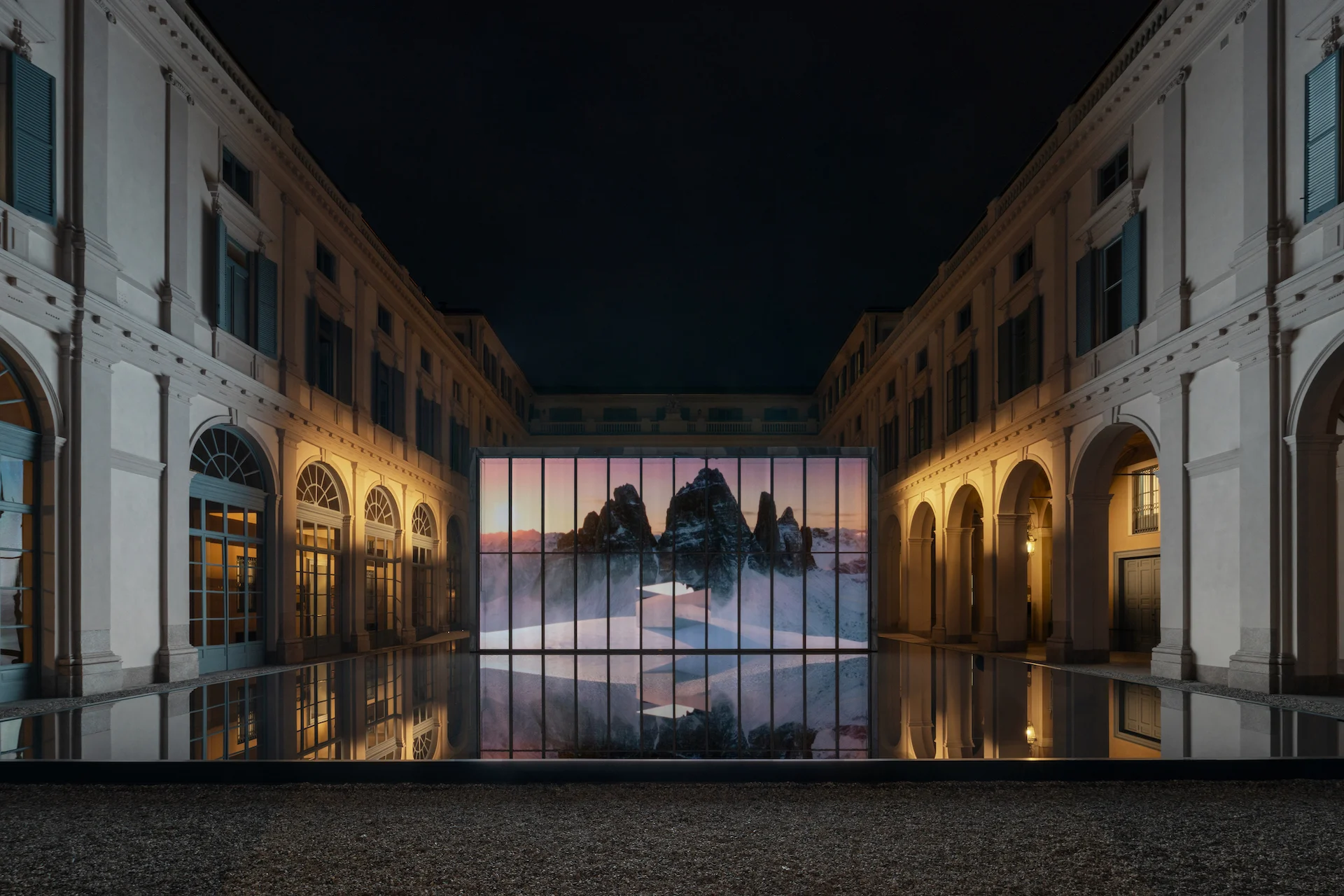 Courtyard at night: glass pavilion glowing with an image of jagged mountain peaks, reflected in the black pool – Samsung House at Palazzo Serbelloni, Milan, Italy, 2026 – interior design