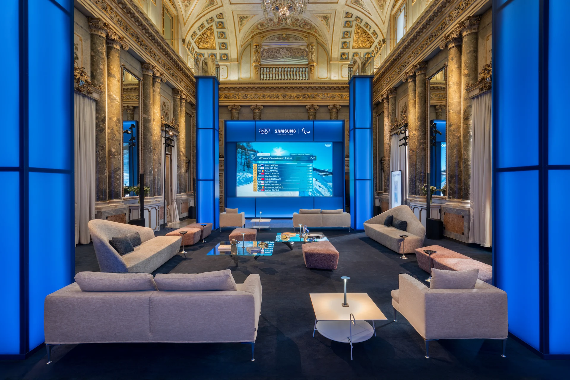 Frescoed palazzo hall with blue light columns, pale sofas and a large screen showing Olympic results – Samsung House at Palazzo Serbelloni, Milan, Italy, 2026 – interior design