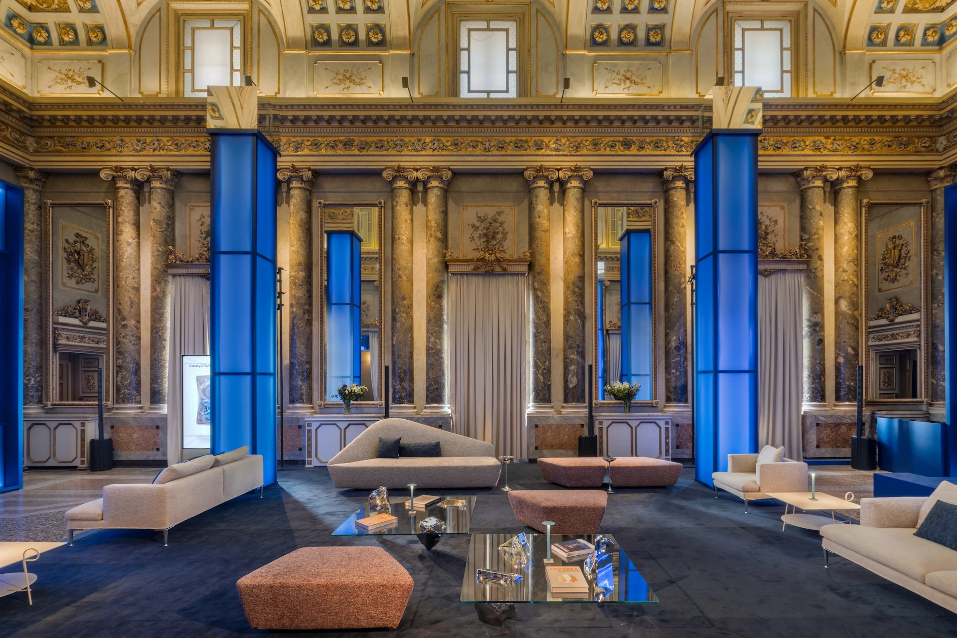 Gilded hall with marble columns and blue-lit pillars, furnished with pale sofas, glass tables and pink ottomans – Samsung House at Palazzo Serbelloni, Milan, Italy, 2026 – interior design