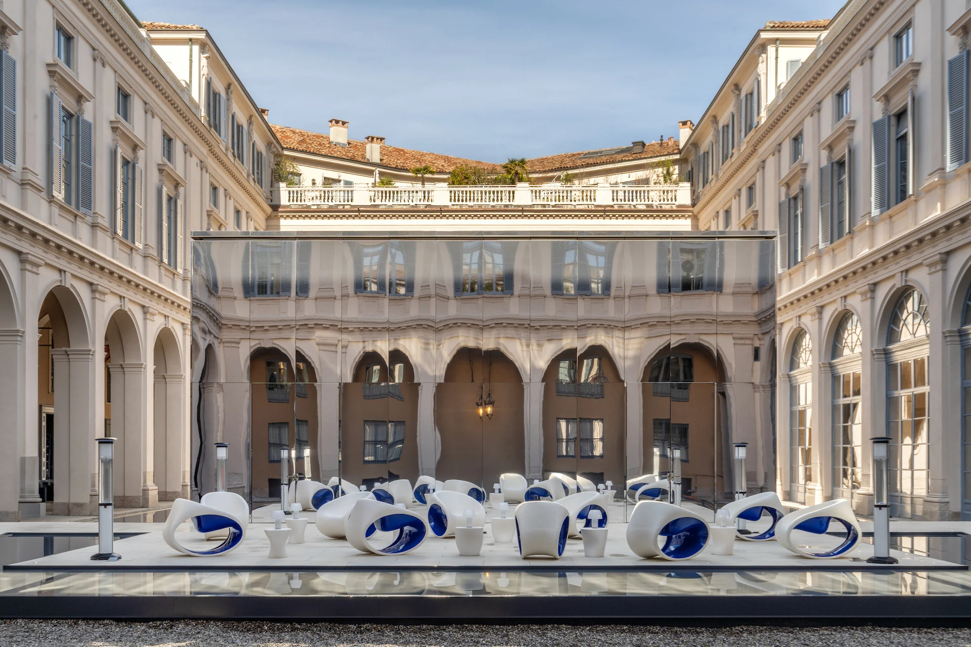 White and blue lounge chairs on a reflective platform in the courtyard, glass pavilion and arcades behind – Samsung House at Palazzo Serbelloni, Milan, Italy, 2026 – interior design