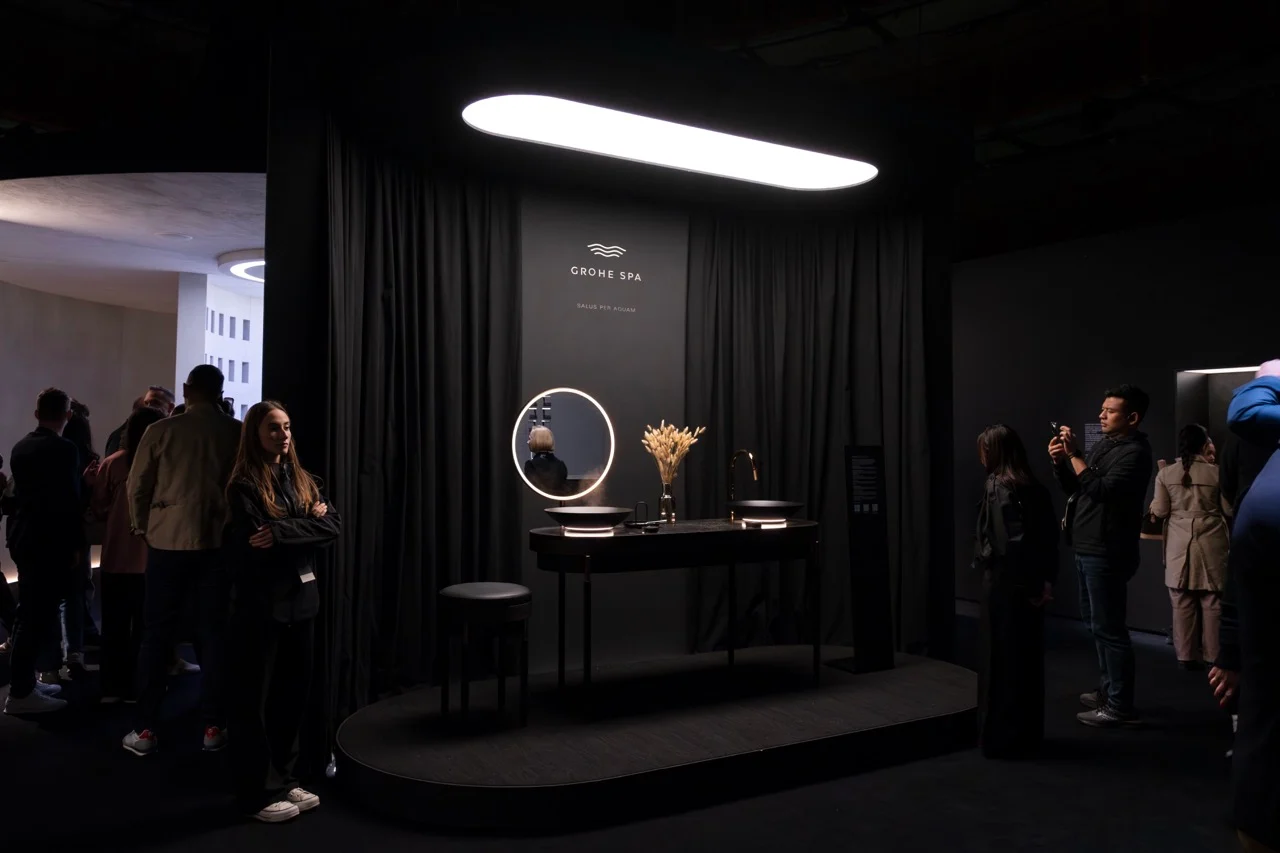 Dark vanity with a round illuminated mirror under an oval light, visitors around the curtained booth – GROHE SPA Aqua Sanctuary at Fuorisalone, Milan, Italy, 2026 – installation design