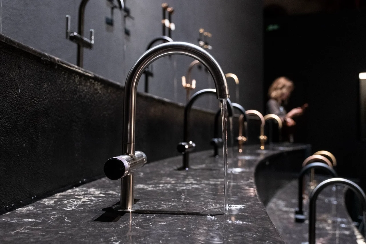 Close-up of a steel tap with running water over a curved dark marble counter lined with taps – GROHE SPA Aqua Sanctuary at Fuorisalone, Milan, Italy, 2026 – installation design
