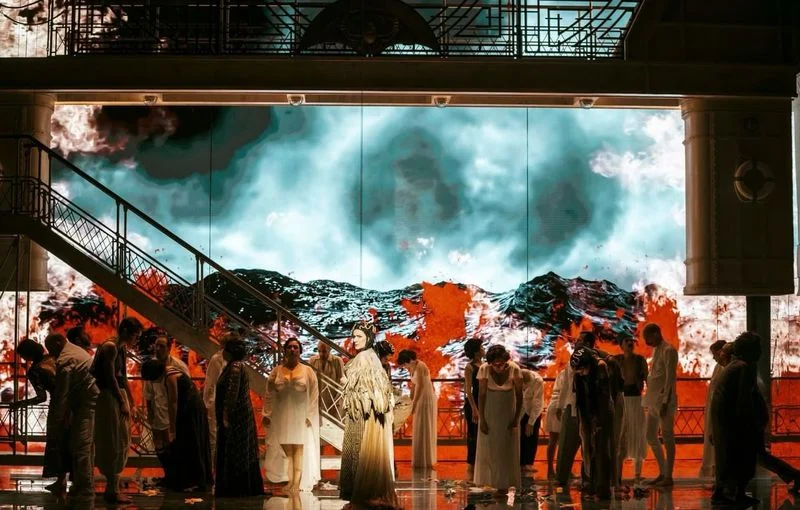 Chorus in pale costumes in front of a projected sea of fire and dark clouds – Giulio Cesare in Egitto at Opéra de Monte-Carlo, Monte Carlo, Monaco, 2025 – set design