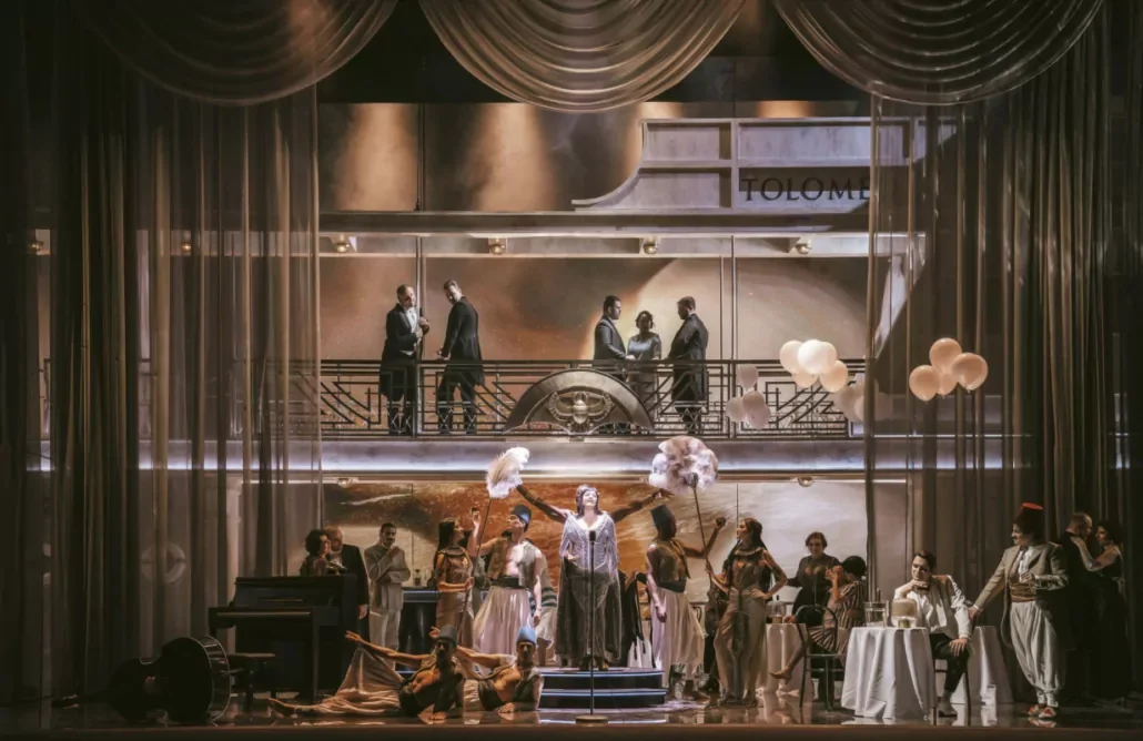Singer on a small stage under draped curtains, with dancers, café tables and men on a balcony above – Giulio Cesare in Egitto at Opéra de Monte-Carlo, Monte Carlo, Monaco, 2025 – set design