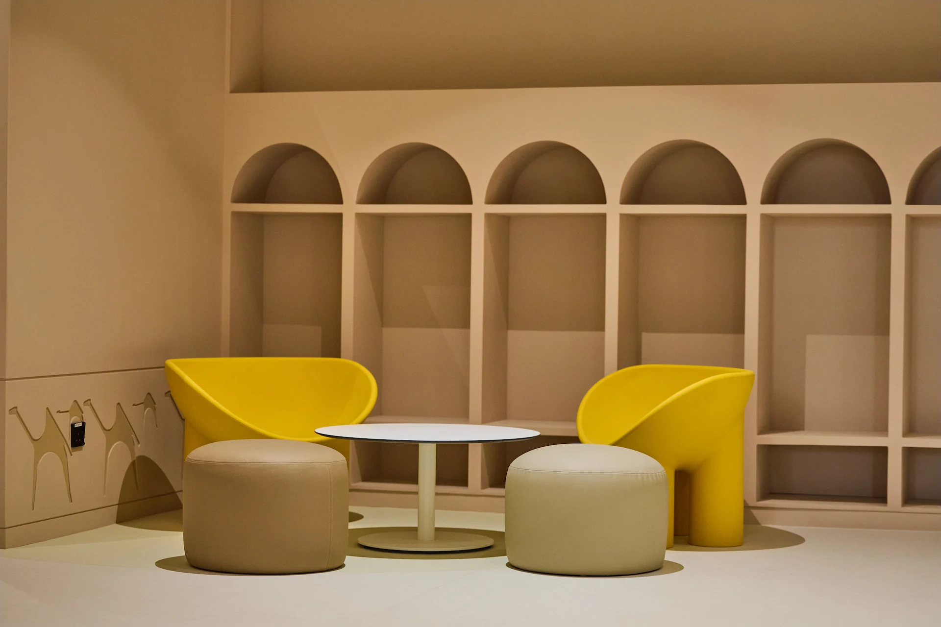 Lounge corner with yellow armchairs and round poufs in front of arched niches in a sand-coloured wall – MOC Headquarters, Diriyah, Saudi Arabia, 2026 – interior design