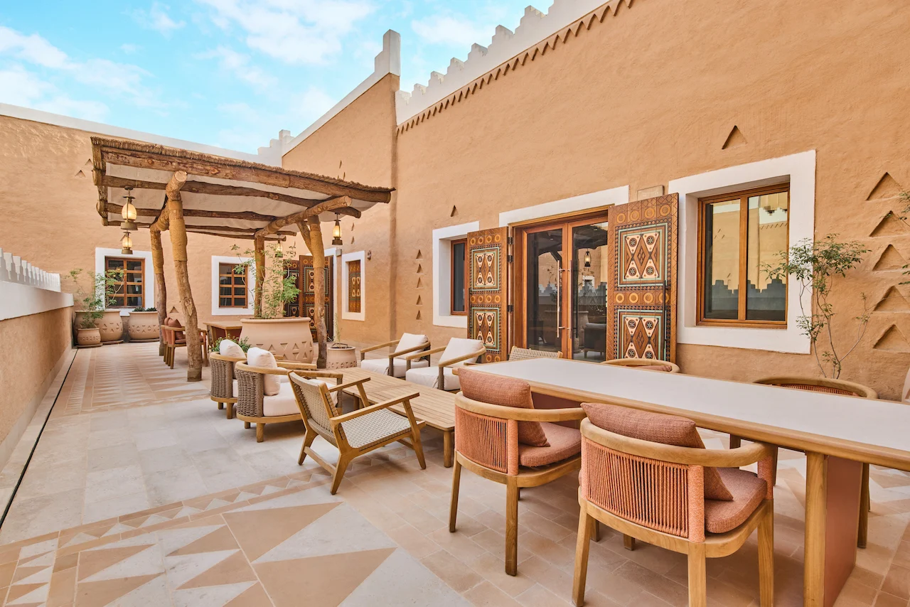 Outdoor terrace with a wooden pergola, dining table and lounge chairs beside earthen walls with painted doors – MOC Headquarters, Diriyah, Saudi Arabia, 2026 – interior design