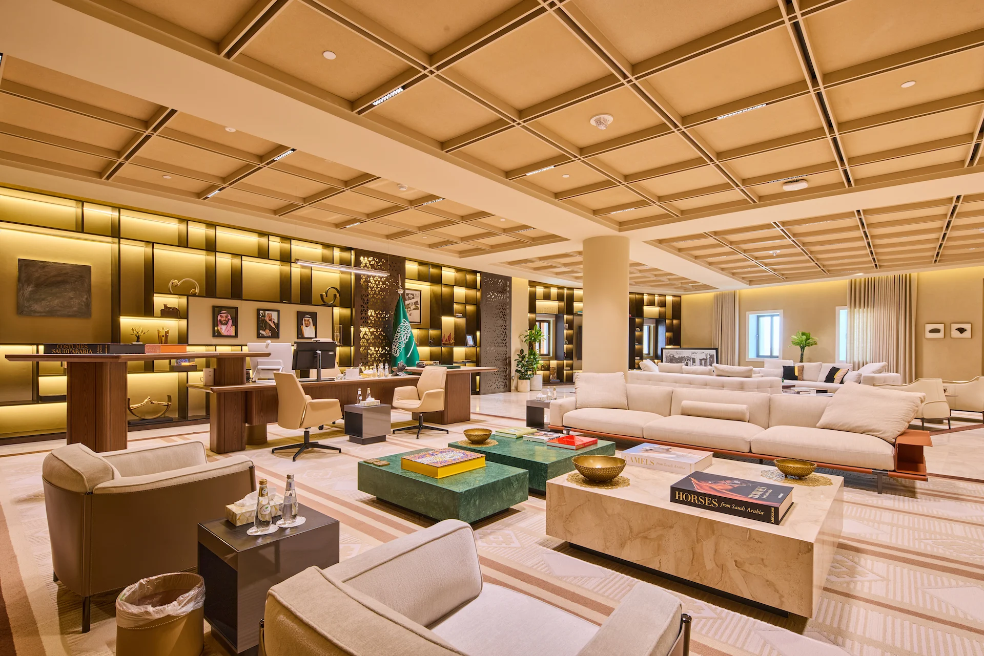 Open-plan executive office and lounge under a coffered ceiling, with backlit shelving, sofas and green marble tables – MOC Headquarters, Diriyah, Saudi Arabia, 2026 – interior design