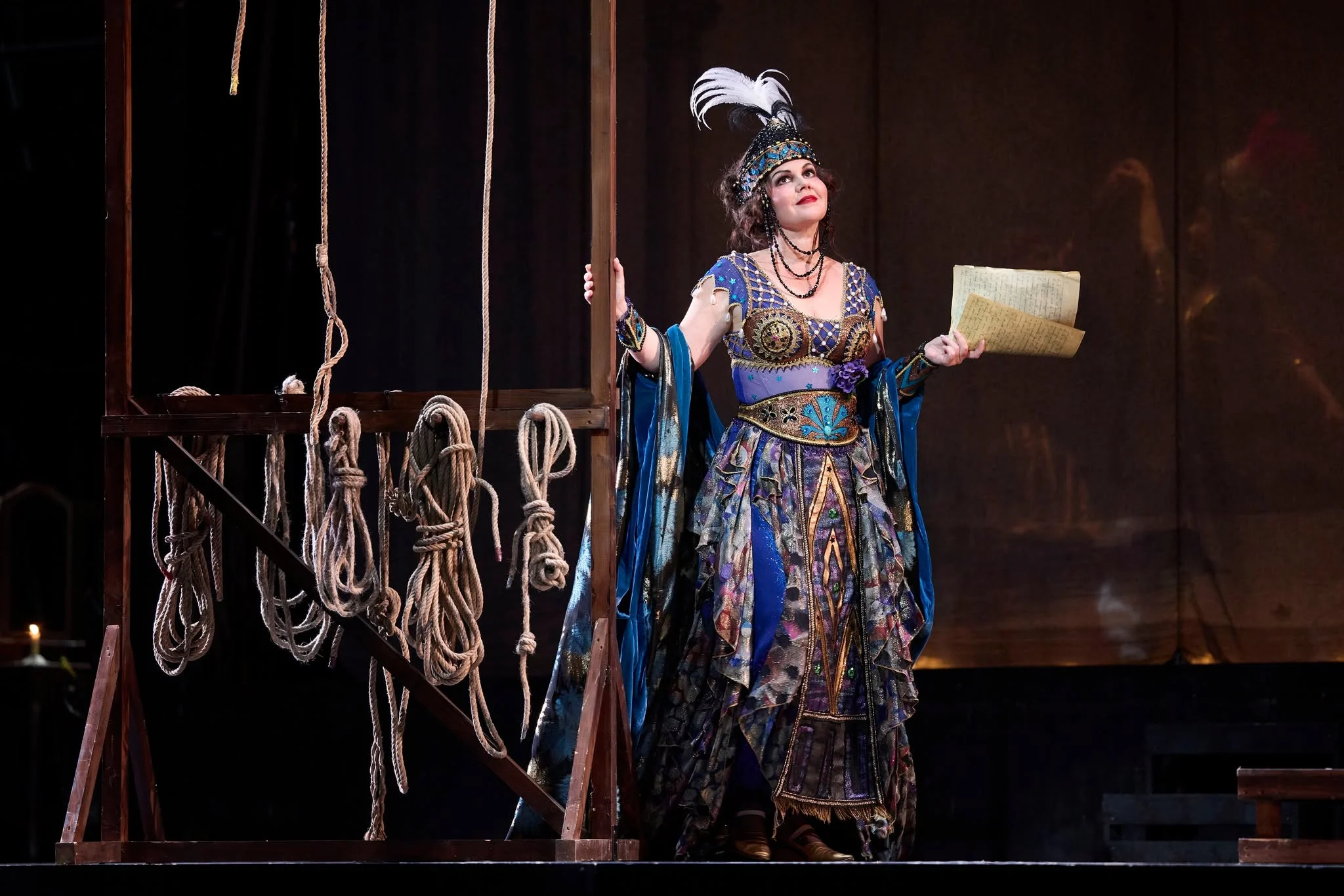 Singer in a jewelled blue costume and feathered headdress holding a letter, beside a frame hung with ropes – Adriana Lecouvreur at Opéra de Monte-Carlo, Monte Carlo, Monaco, 2026 – set design
