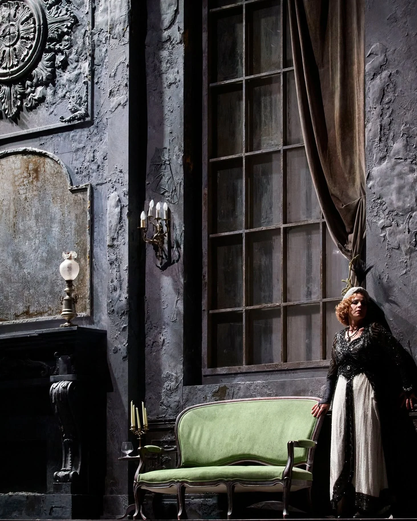 Singer in black lace beside a green settee, in a weathered grey salon with a tall window – Adriana Lecouvreur at Opéra de Monte-Carlo, Monte Carlo, Monaco, 2026 – set design