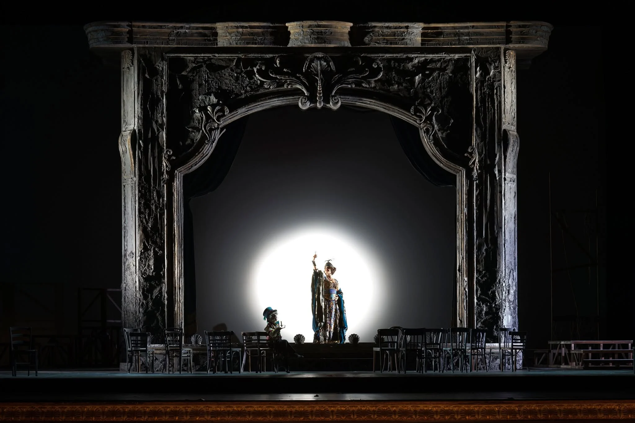 Singer in a white spotlight at the centre of a carved silver portal, chairs lined up on the dark stage – Adriana Lecouvreur at Opéra de Monte-Carlo, Monte Carlo, Monaco, 2026 – set design