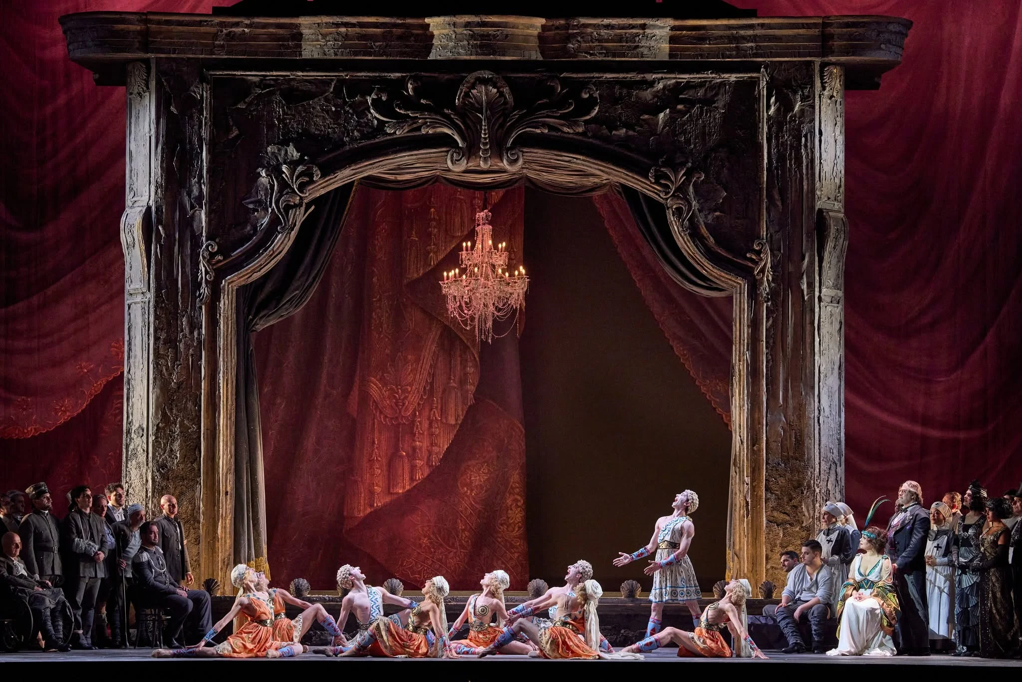 Dancers in orange costumes beneath a chandelier, framed by the carved portal and red velvet drapes – Adriana Lecouvreur at Opéra de Monte-Carlo, Monte Carlo, Monaco, 2026 – set design