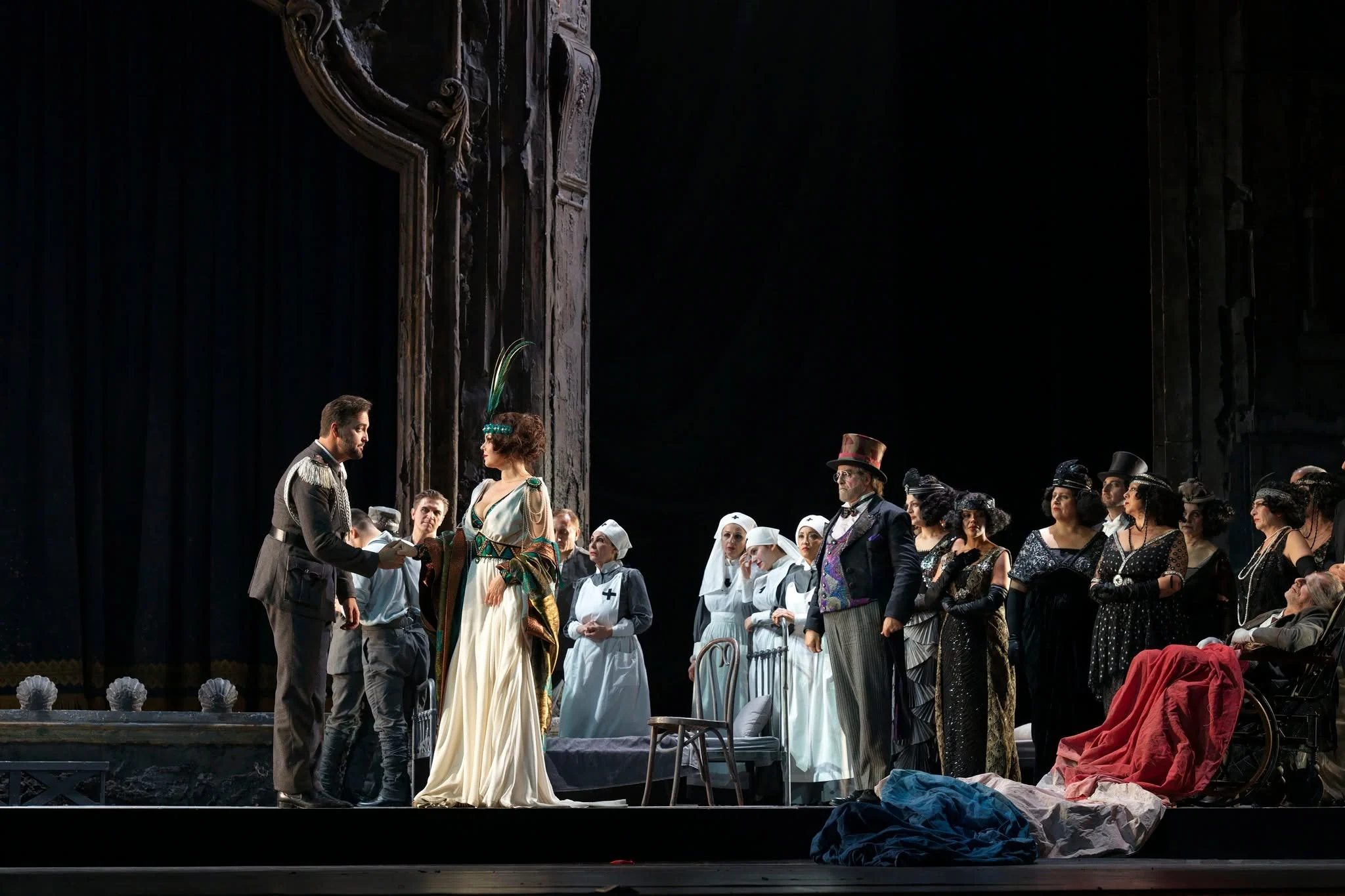 Soloists and chorus in period costume with nurses in white, gathered beside the carved portal – Adriana Lecouvreur at Opéra de Monte-Carlo, Monte Carlo, Monaco, 2026 – set design
