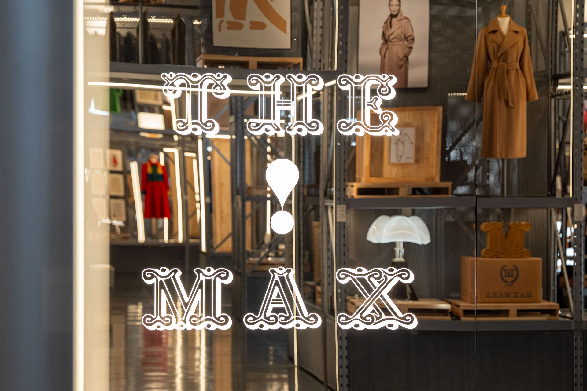Exhibition title The Max! on a glass door, with Max Mara coats displayed on metal shelving behind – Max Mara The Max! at Long Museum, Shanghai, China, 2026 – exhibition design