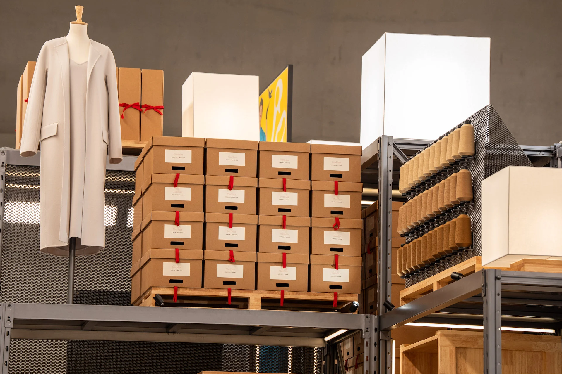 Pallets of labelled archive boxes tied with red ribbon beside a long ivory coat on a mannequin – Max Mara The Max! at Long Museum, Shanghai, China, 2026 – exhibition design