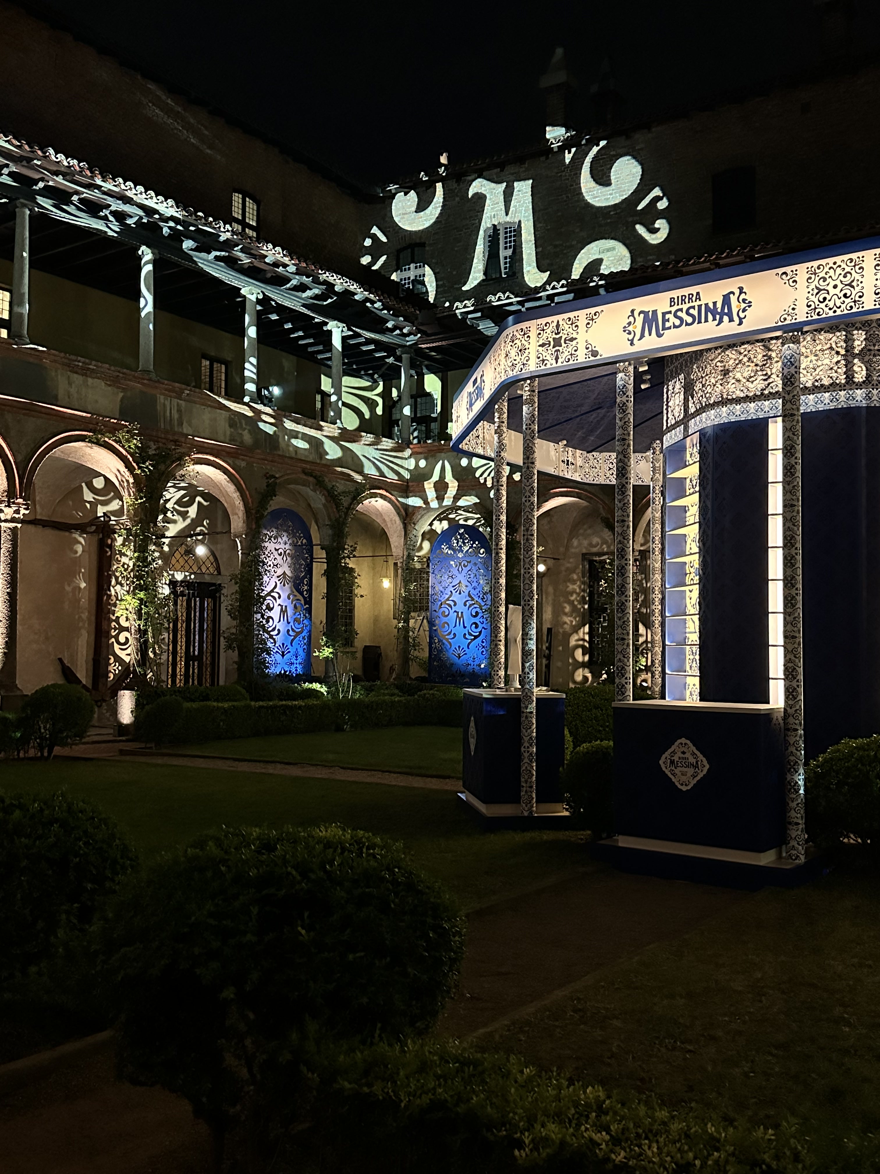 Courtyard at night with a Birra Messina kiosk and patterned blue projections on the arcaded façade – Birra Messina Bar Meraviglia at Fuorisalone, Milan, Italy, 2024 – event design